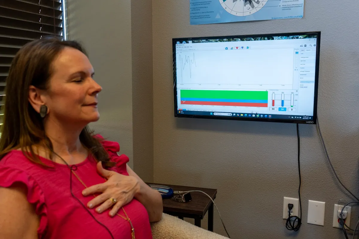 A woman sitting with her eyes closed and a hand on her chest, watching coloured coherence bars on a wall-mounted screen.